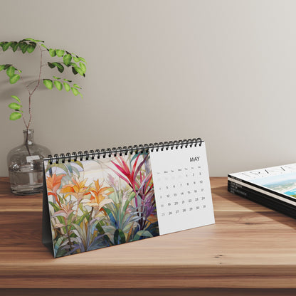 A 2025 Desk Calendar - Tropical Flowers set to May, showcasing vibrant botanical artwork with a tropical flower theme. It is placed on a wooden surface beside a small plant in a glass vase and a stack of closed magazines. Made in the USA.