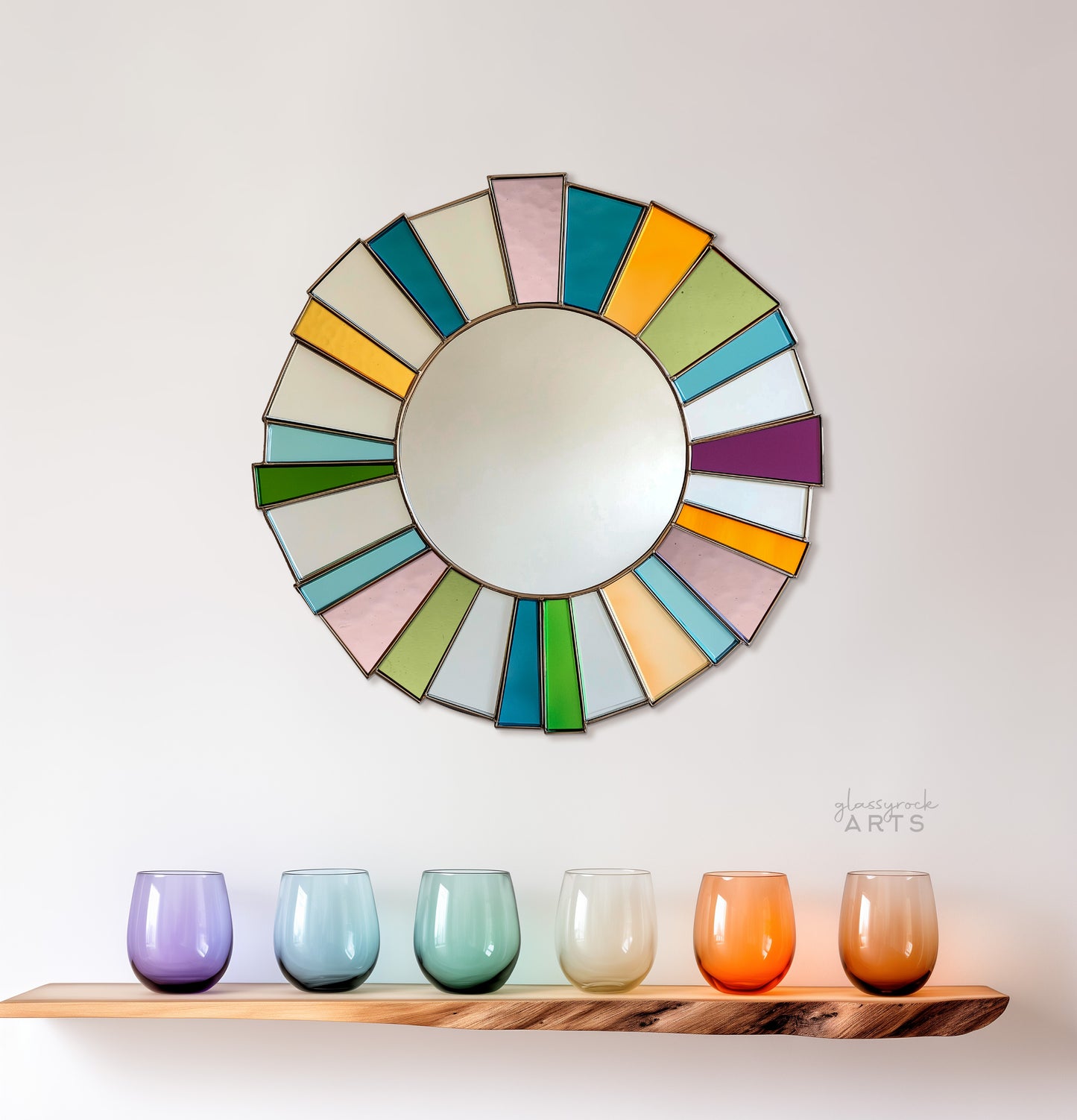 A round mirror surrounded by bright multicolored pieces of stained glass radiating from the edges of the mirror. It hangs on a wall with colorful glasses on a natural wood shelf below.