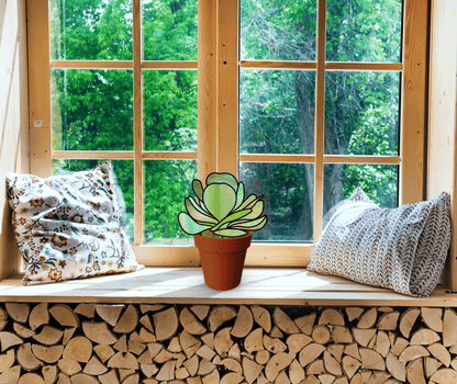 A potted plant sits on a wooden windowsill, flanked by two cushions with the Stained Glass Succulent Stems Pattern Pack of 4. The window reveals a lush green view outside, while logs are neatly stacked beneath the sill.