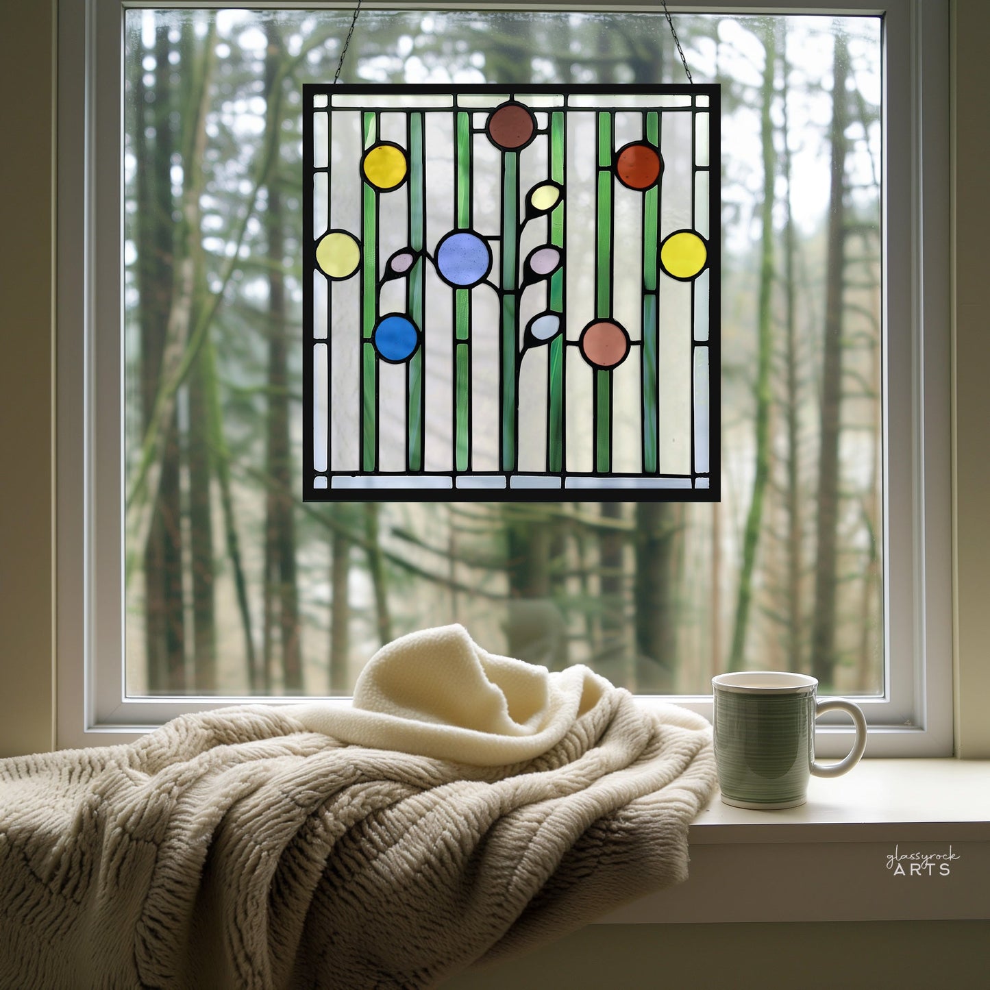 An Abstract Garden stained glass panel with colorful geometric flowers adorns the window. A beige blanket and a green mug rest on the windowsill, while blurred slender trees form an outdoor backdrop.