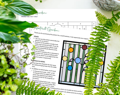 The Abstract Garden Stained Glass Flowers Pattern guide lies on a table among lush plants, featuring instructions and a vibrant geometric design with circular and rectangular shapes, reminiscent of abstract stained glass templates.