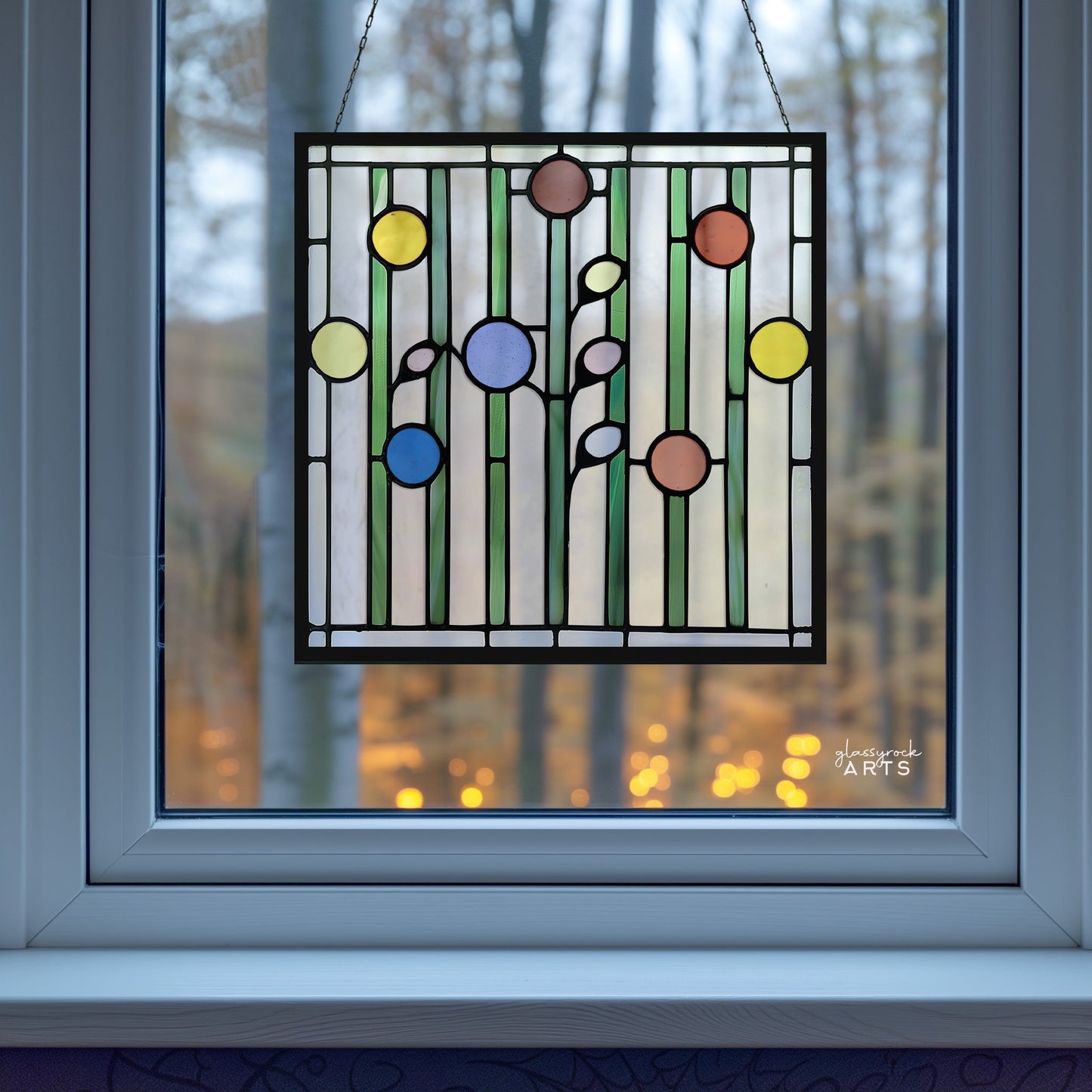 The Abstract Garden Stained Glass Flowers Pattern panel hangs in a window, highlighting an abstract design with vertical lines and circles in red, blue, green, and yellow. Outside, a blurred view shows tall trees and warm-toned bokeh lights.