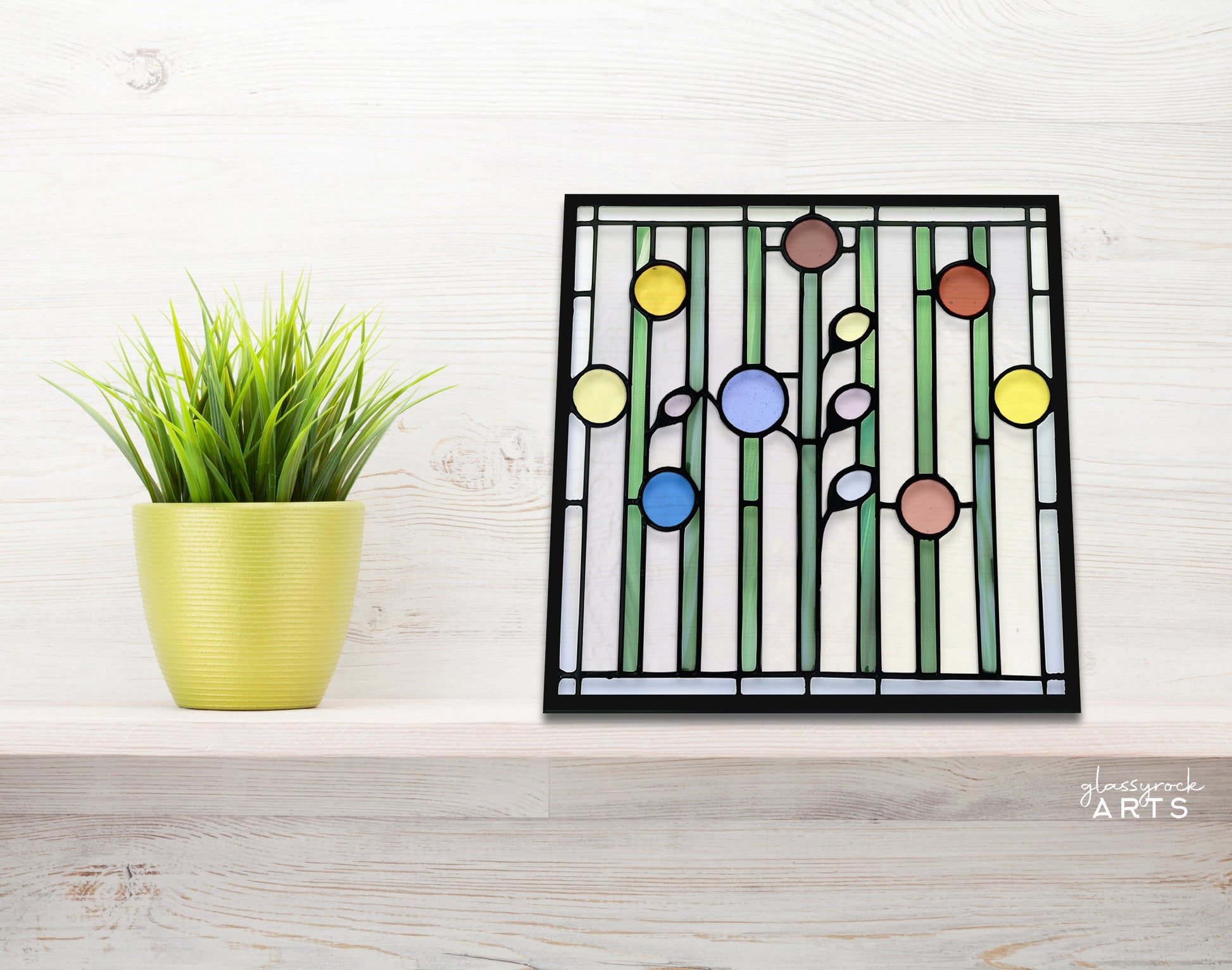 The Abstract Garden Stained Glass Flowers Pattern is displayed on a wooden shelf beside a potted plant, showcasing colorful circular designs framed in black.