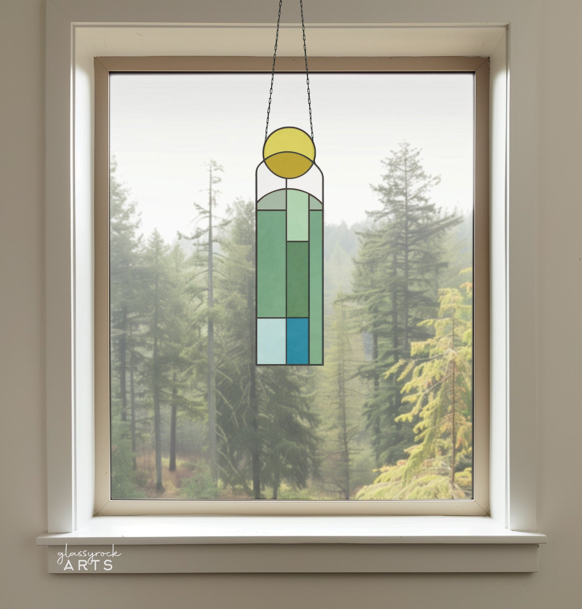 A window overlooks a foggy forest of tall evergreens. Hanging in front is the Abstract Sunset Stained Glass Pattern sun catcher, featuring geometric green, yellow, and blue tones that craft an abstract digital design against the misty backdrop.