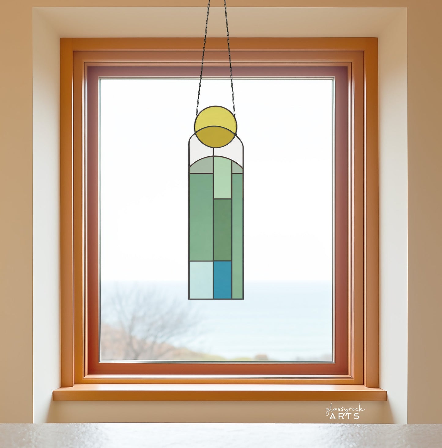 The Abstract Sunset Stained Glass Pattern, with its geometric shapes in green, blue, and yellow, is framed in wood against a window. It evokes the feel of an abstract sunset with a blurred view of trees and the sea outside, suggesting tranquil coastal vibes.