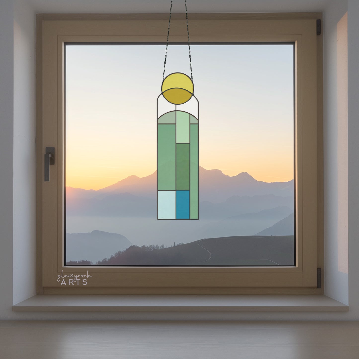 An abstract sun and landscape stained glass panel in colors of yellow, white, blue. The panel is rectangular and tall and it hangs in a window with mountains and water behind.