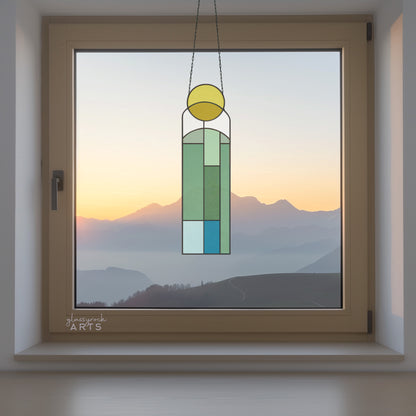 An abstract sun and landscape stained glass panel in colors of yellow, white, blue. The panel is rectangular and tall and it hangs in a window with mountains and water behind.