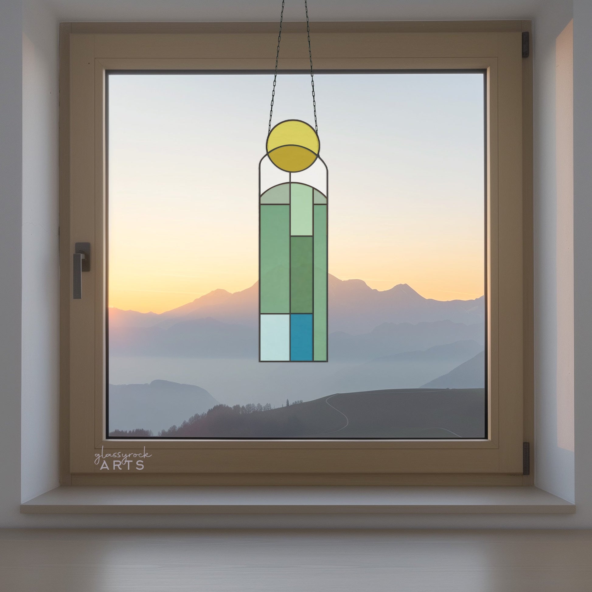 Displayed in the window is the Abstract Sunset Stained Glass Pattern, a geometric-style sun catcher featuring blue, green, and yellow stained glass. It frames a serene mountain landscape with an abstract sunset motif in soft orange and pink hues.