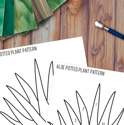 The Beginner Aloe Potted Plant Stained Glass Pattern sheets lie on a wooden surface, paired with a craft knife and green gradient paper sheets, creating a welcoming workspace for DIY projects.