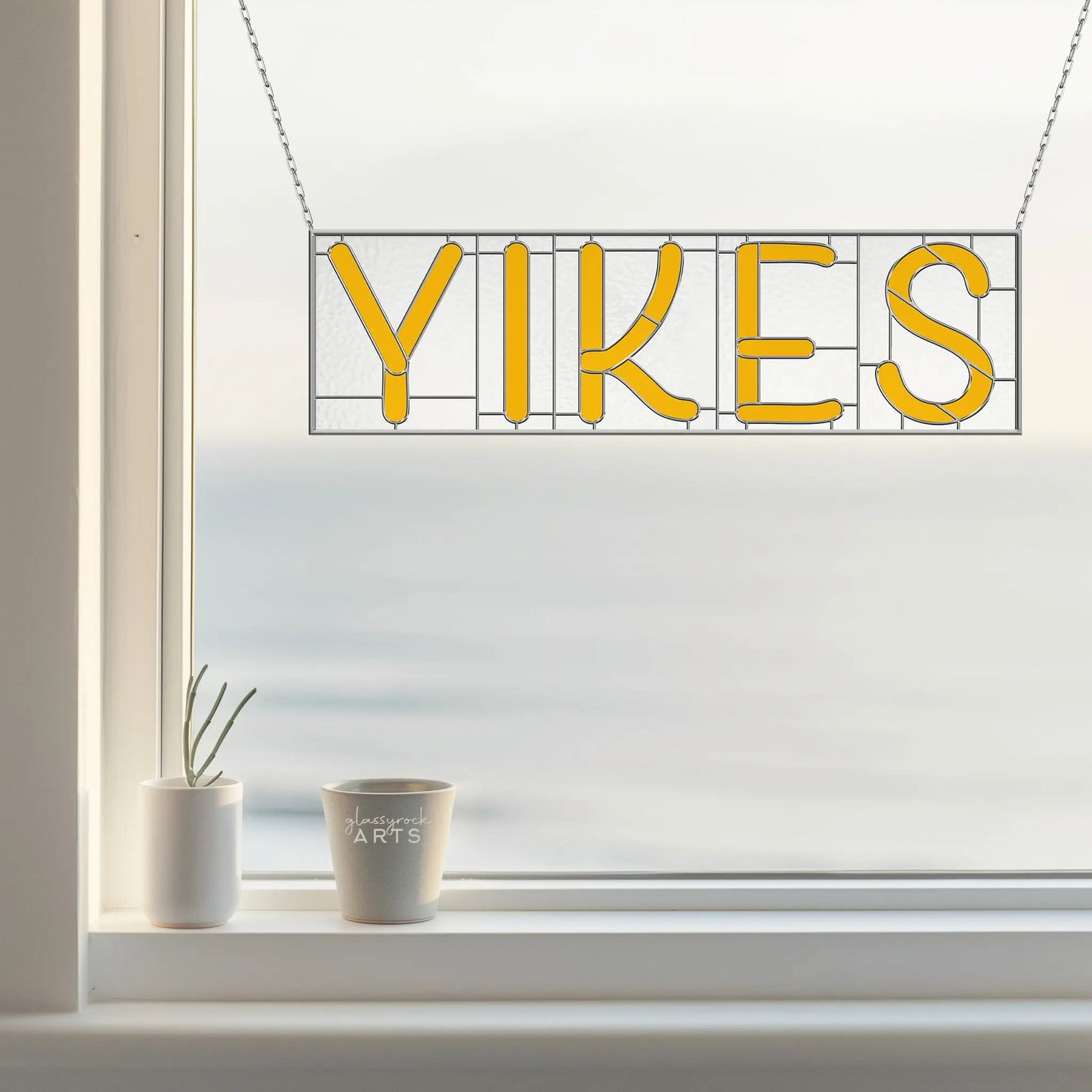 A window is decorated with a stained glass suncatcher from the Stained Glass Alphabet Pattern Pack, with a vibrant yellow YIKES sign hanging. Below, a small plant and a mug labeled LAGOON ARTS sit on the sill, against a backdrop of blurred serenity outside.