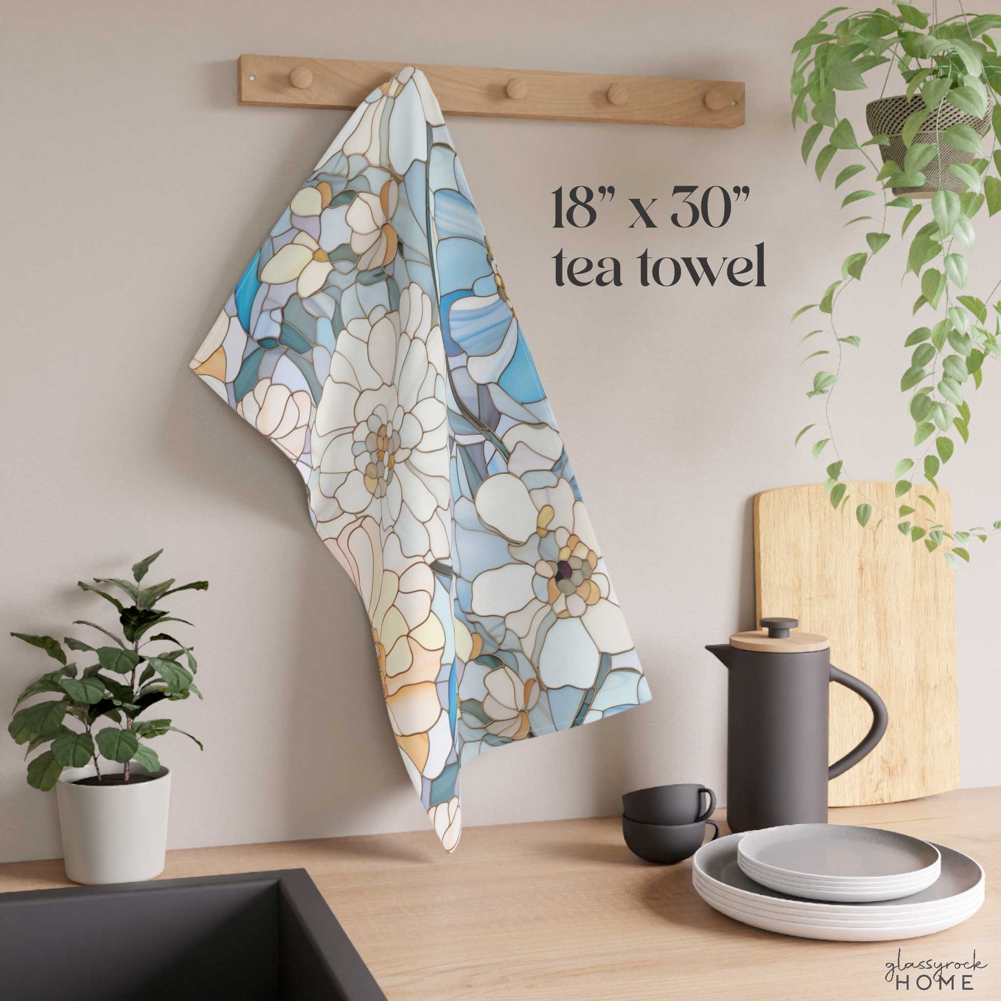 A Stained Glass Flowers Kitchen Towel measuring 18"x30" is hanging on a wooden rack in a kitchen, enhancing the space with a potted plant, kettle, cup, plates, cutting boards, and a sink visible. This charming towel makes for an ideal housewarming gift. Text reads "18" x 30" tea towel".