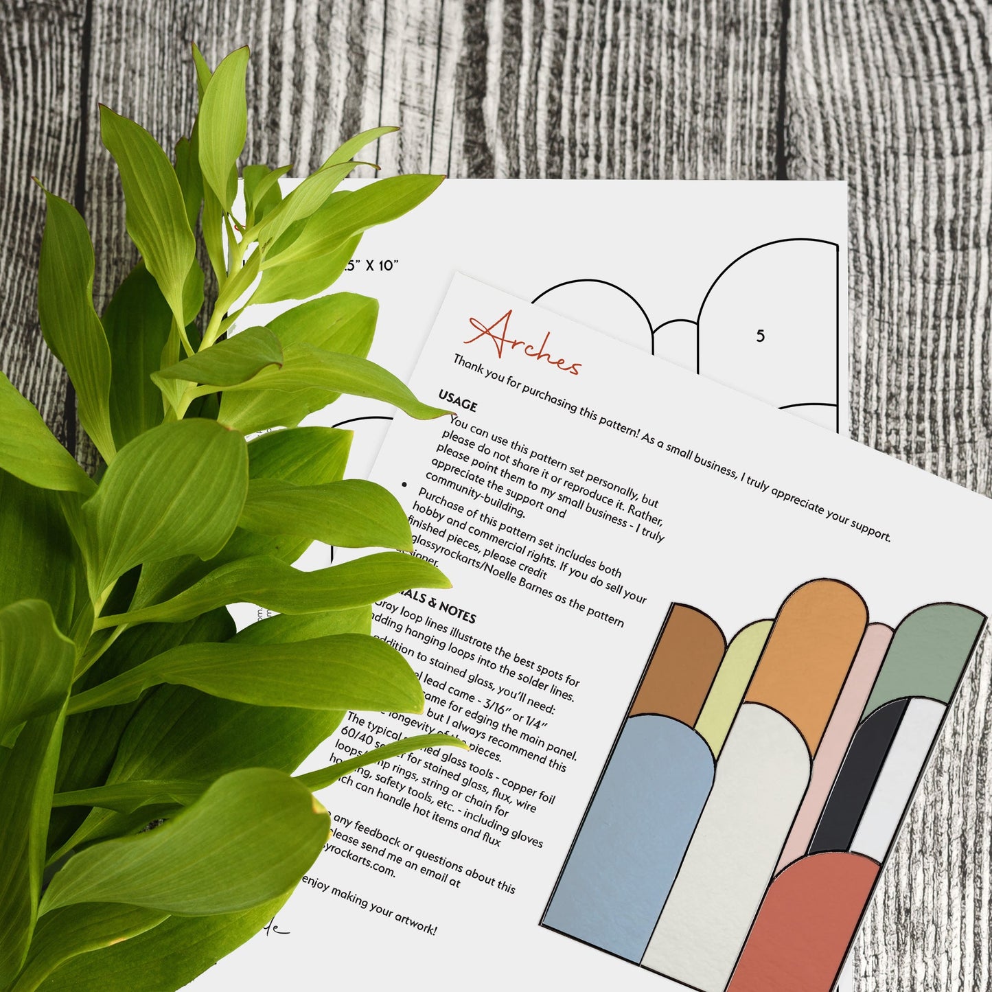 A leafy plant is next to two sheets of paper on a wooden surface. One sheet says Arches above colorful geometric designs from the Retro Arches Stained Glass Pattern collection, with text filling the rest.