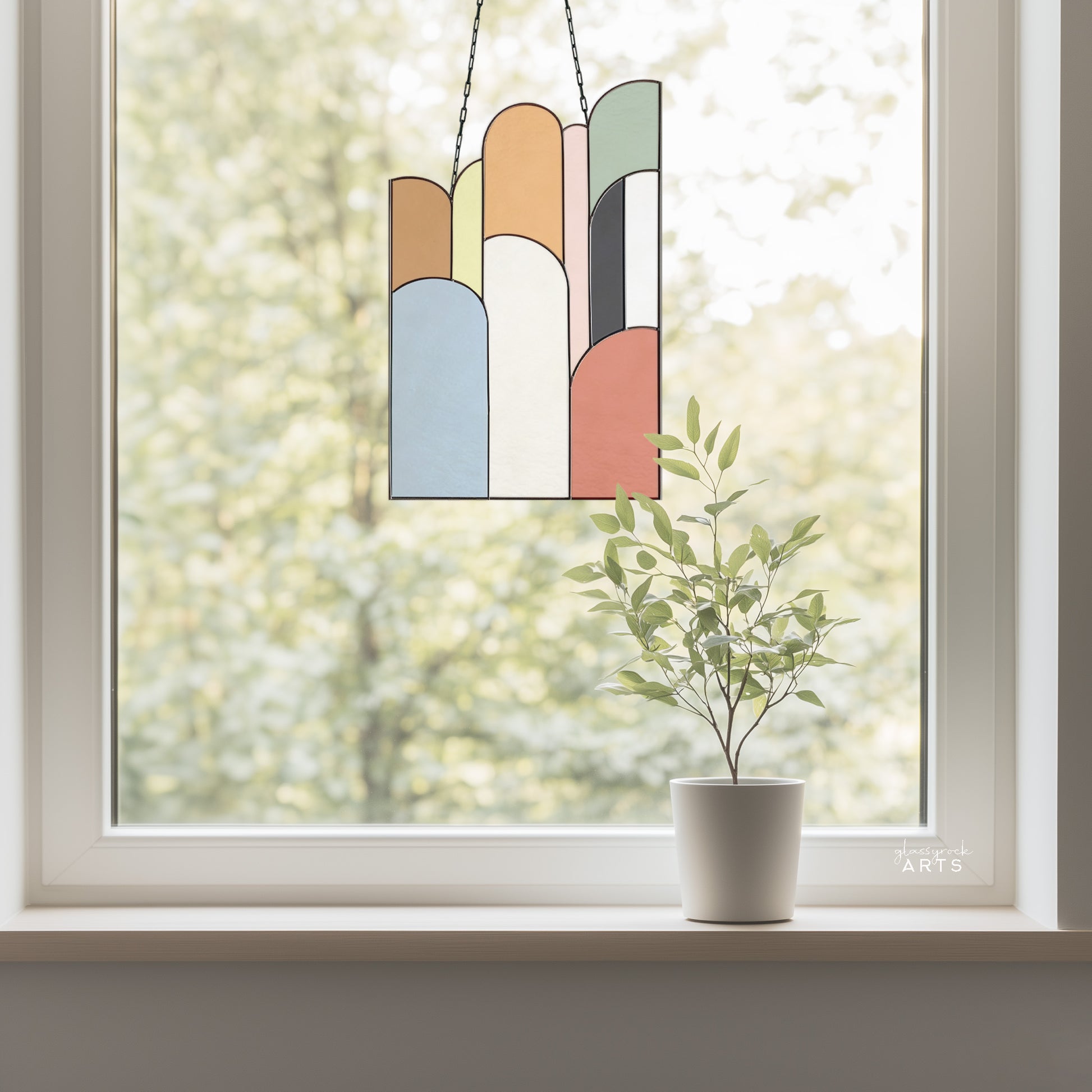 An abstract stained glass panel with ascending arch shapes layered on top of one another in multicolor hues. The panel is rectangular and tall. It hangs in a window with trees behind it.