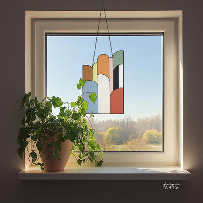 An abstract stained glass panel with ascending arch shapes layered on top of one another in multicolor hues. The panel is rectangular and tall. It hangs in a window with trees behind it.