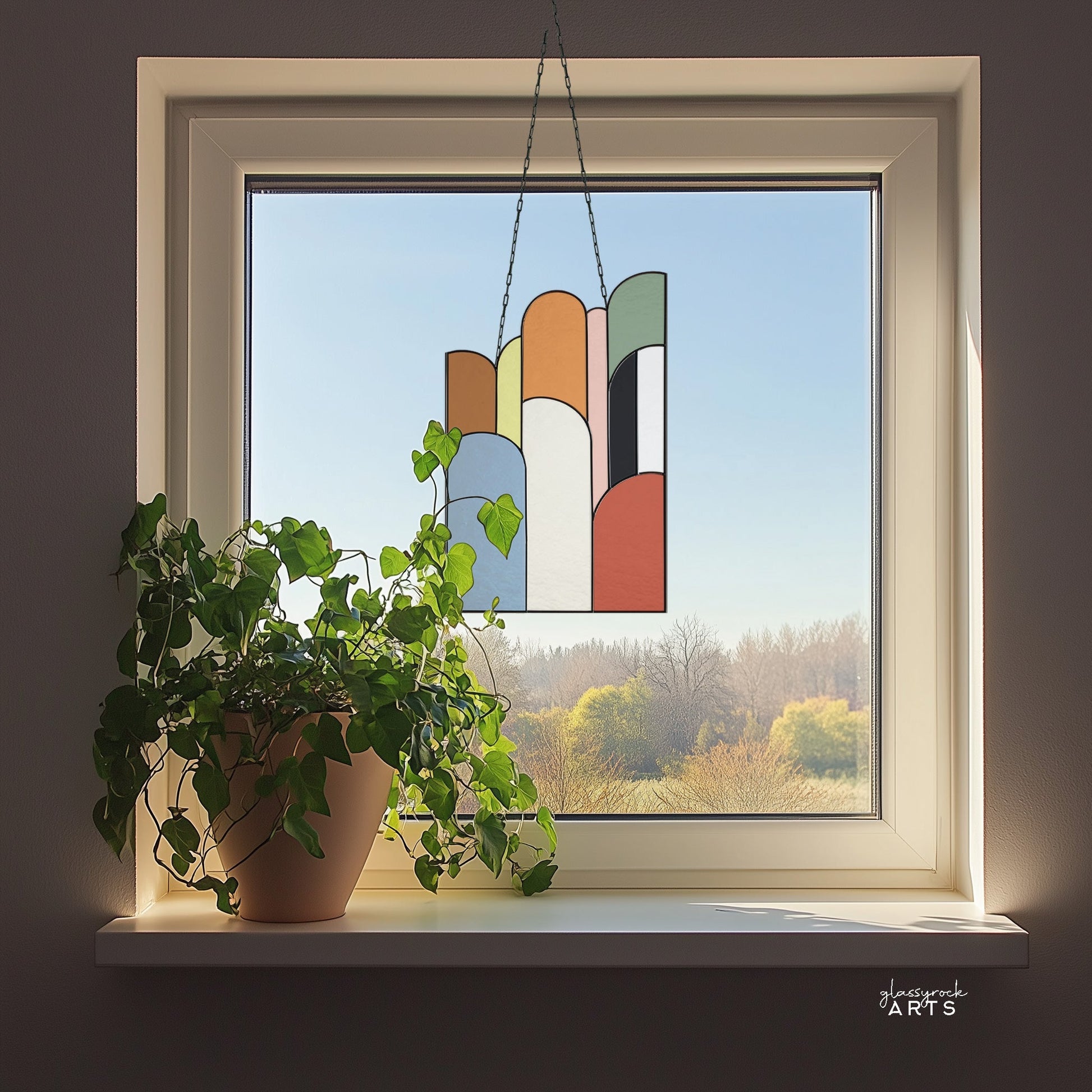 The Retro Arches Stained Glass Pattern decorates a bright window with geometric designs, framing a tranquil view of trees. A green-leaved potted plant enhances the natural beauty of this artistic setup.