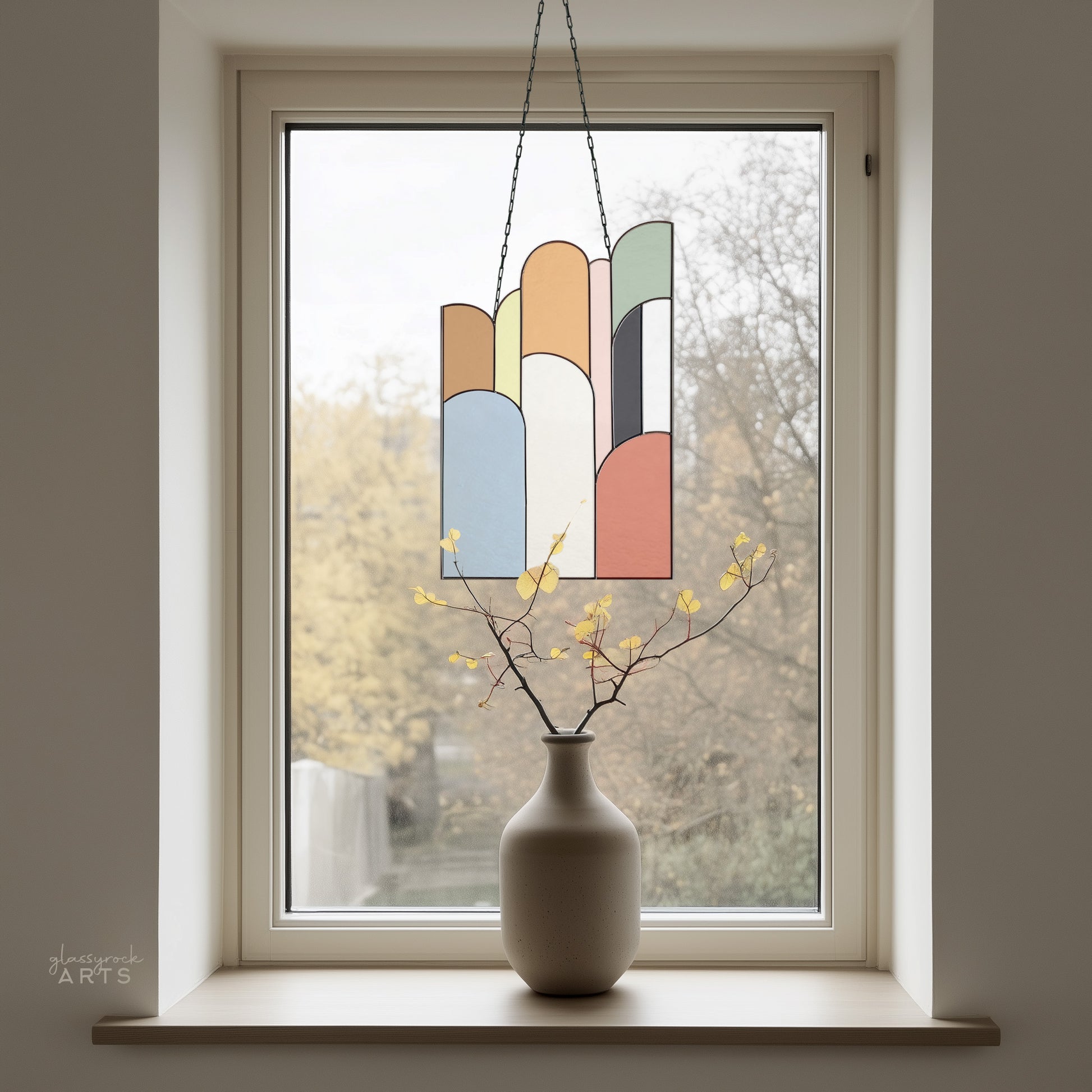 An abstract stained glass panel with ascending arch shapes layered on top of one another in multicolor hues. The panel is rectangular and tall. It hangs in a window with trees behind it and a vase in front.