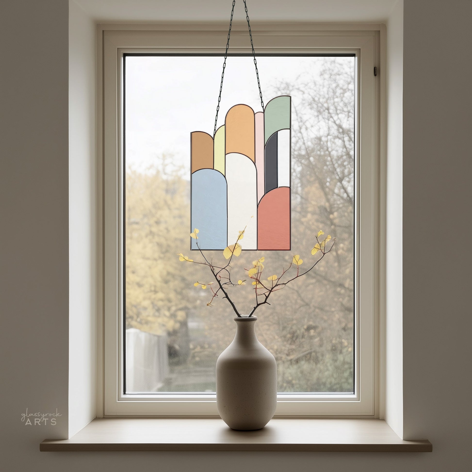 A minimalist window display presents a vase with yellow budding branches and the Retro Arches Stained Glass Pattern hanging above, creating a stunning sun catcher effect. The window offers a soft-focus view of a light, neutral-toned landscape.