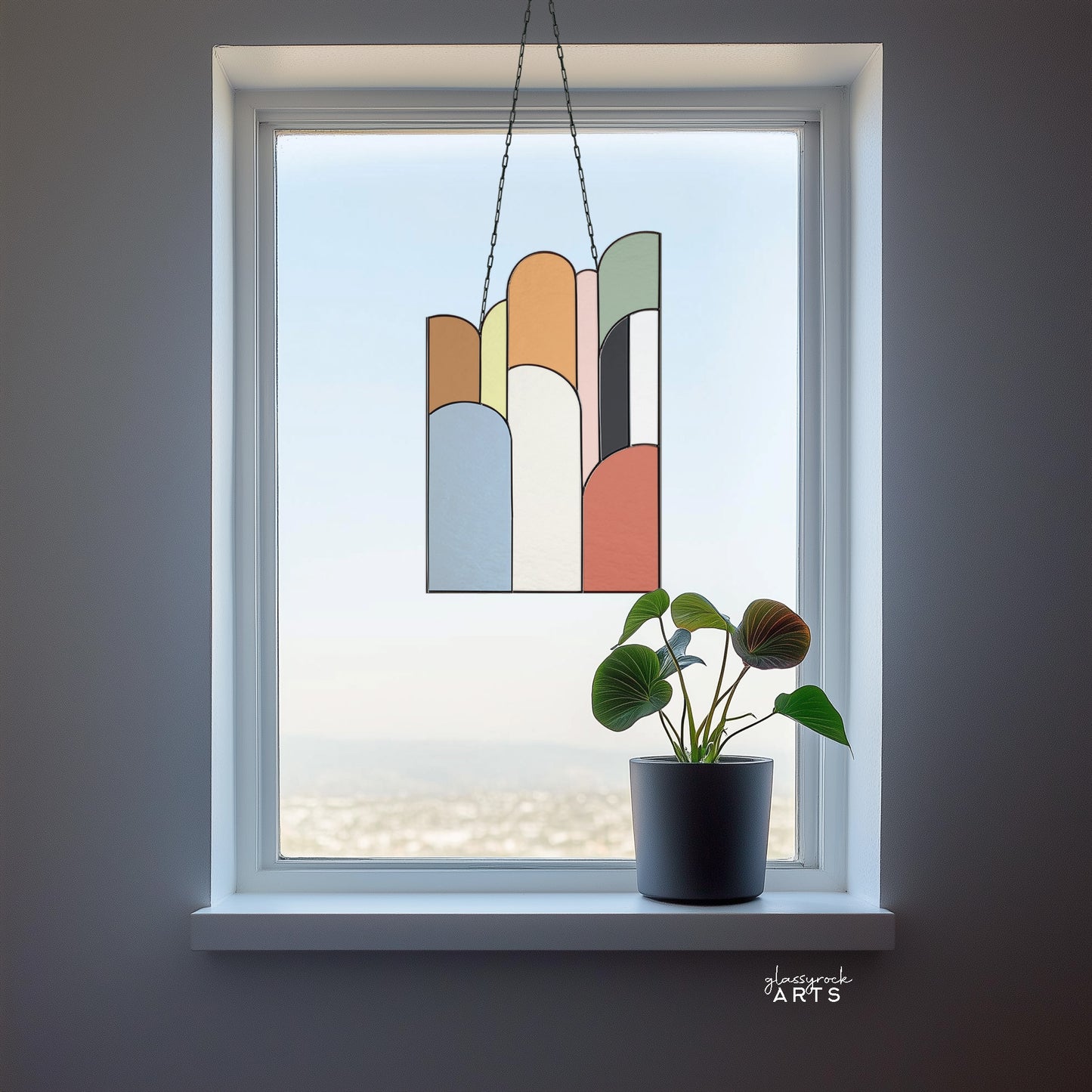 An abstract stained glass panel with ascending arch shapes layered on top of one another in multicolor hues. The panel is rectangular and tall. It hangs in a window with a city landscape behind it.