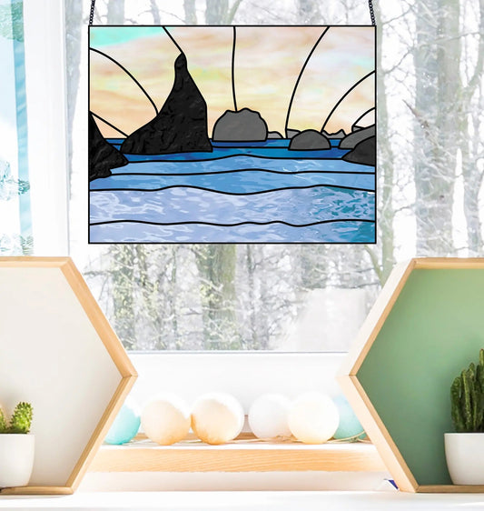 The Oregon Seastack Rocks Stained Glass Pattern adorns a window, depicting a seascape with rocks and sunset. Below, hexagonal shelves display décor: white balls on the left and a cactus with white accents on the right. Snow-covered trees are visible outside.