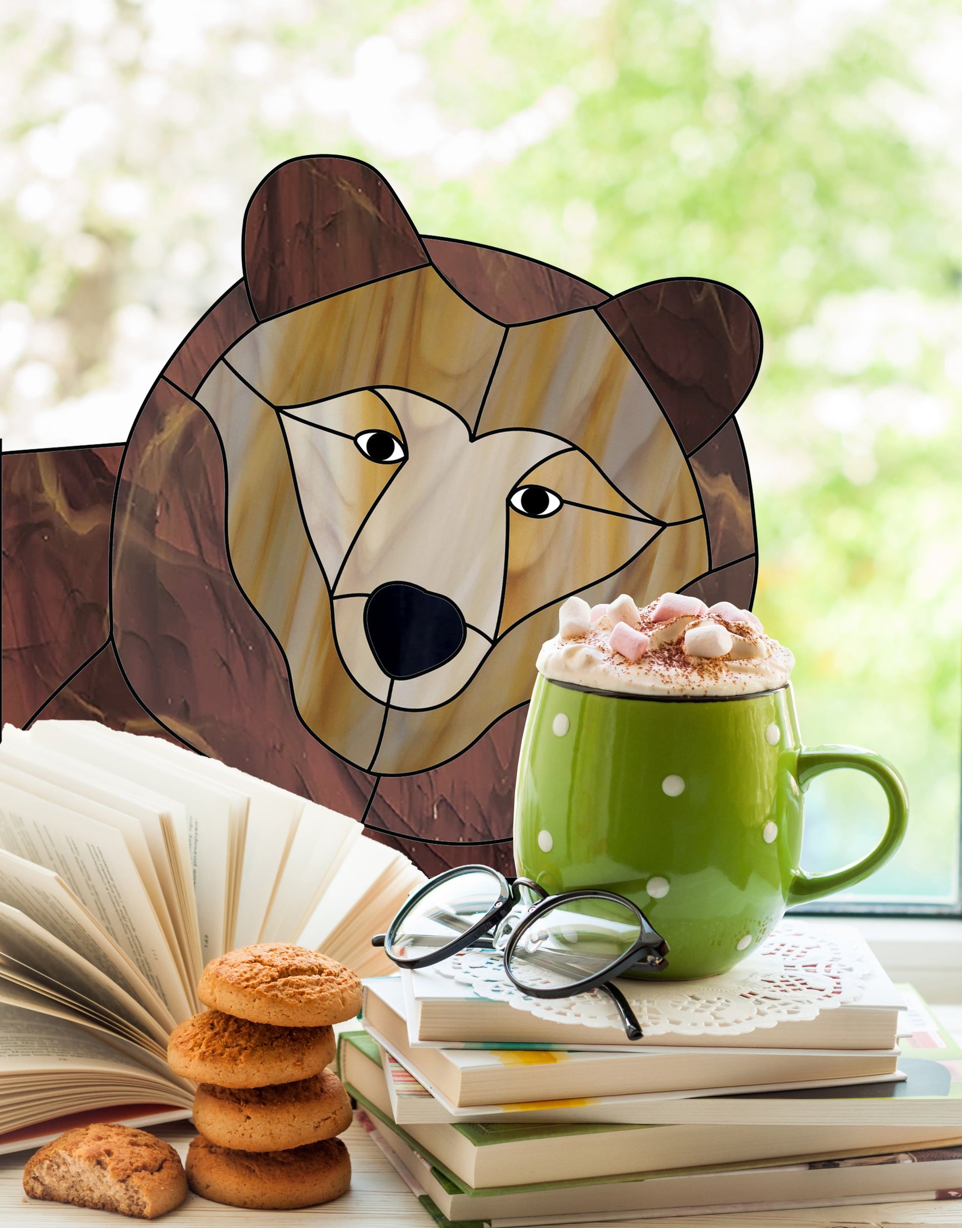 A bear illustration is visible through a window next to an open book, a stack of cookies, a pair of glasses, and a green mug filled with a hot beverage topped with marshmallows.