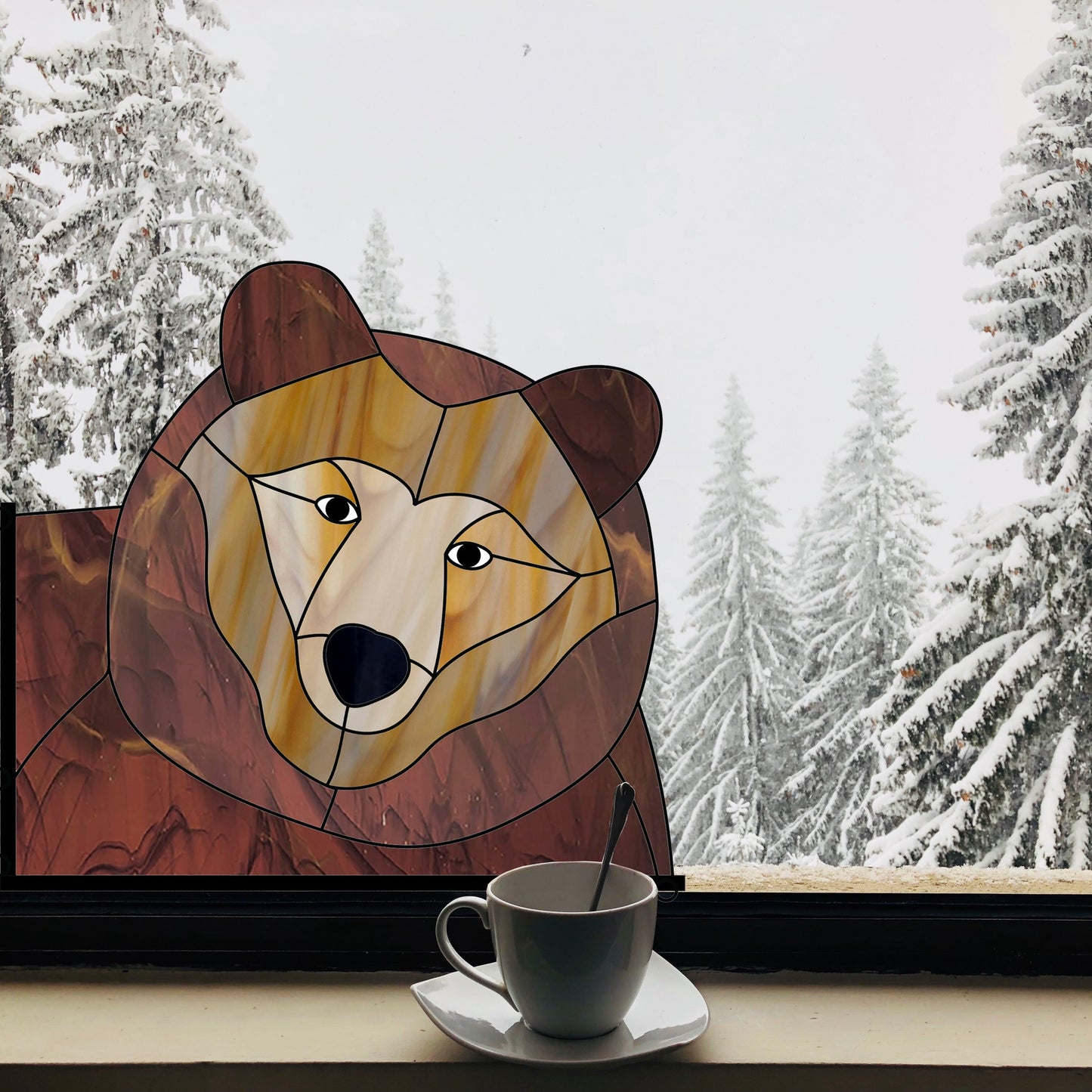 A stained glass bear is mounted on a window with a snowy forest view behind it. A cup of tea on a saucer sits on the window sill.