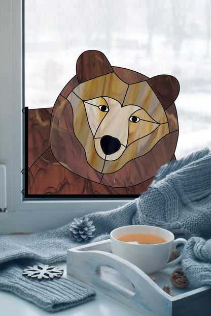 A stained glass bear decoration is placed on a window. In front of the window, there is a tray with a cup of tea, pine cones, and a knitted scarf, with a snowy landscape in the background.