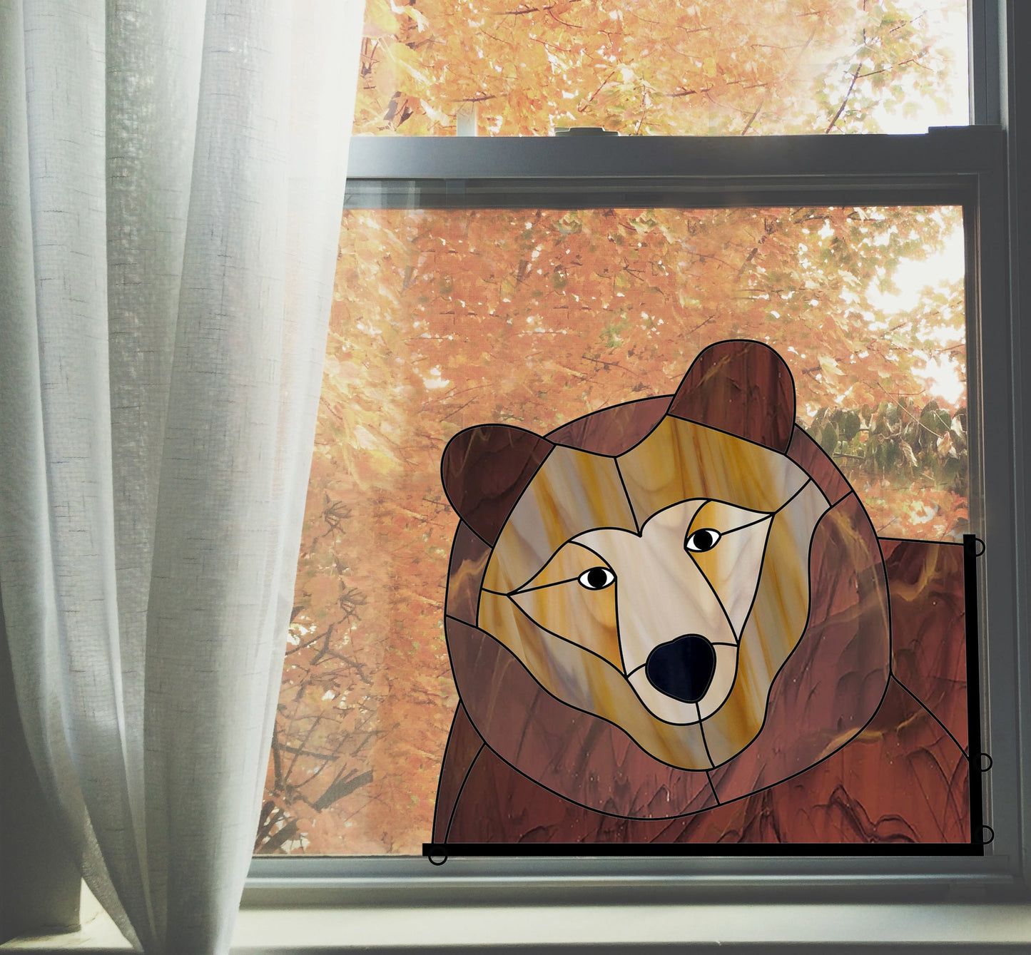 A stained glass artwork depicting a bear is hung on a window, with fall foliage visible outside. A white curtain partially covers the window.