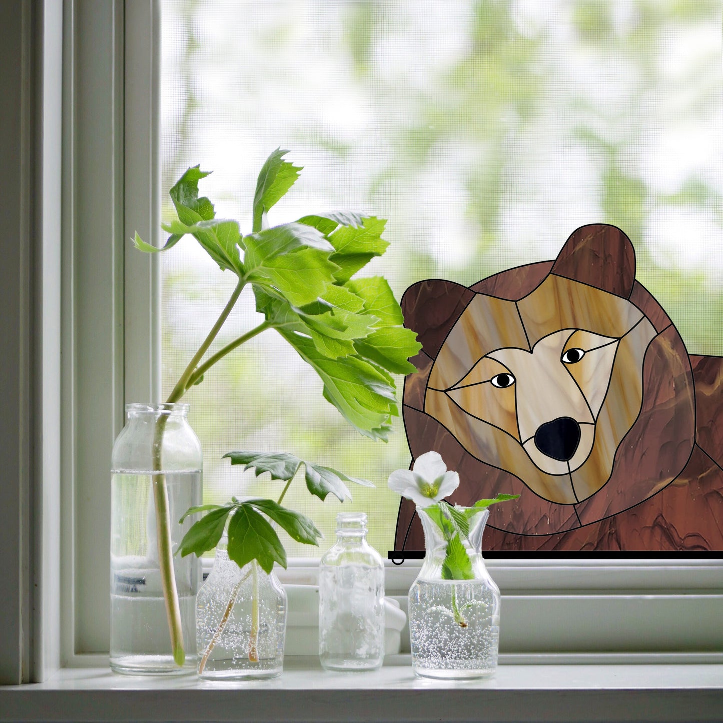 A stained glass bear decoration is attached to a white window frame, next to three glass bottles containing green plant cuttings.