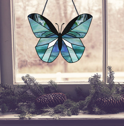 A stained glass butterfly, crafted using the Beginner Butterfly Stained Glass Pattern in blue and green hues, hangs in a window, while pine branches and cones on the windowsill add a natural and cozy touch.