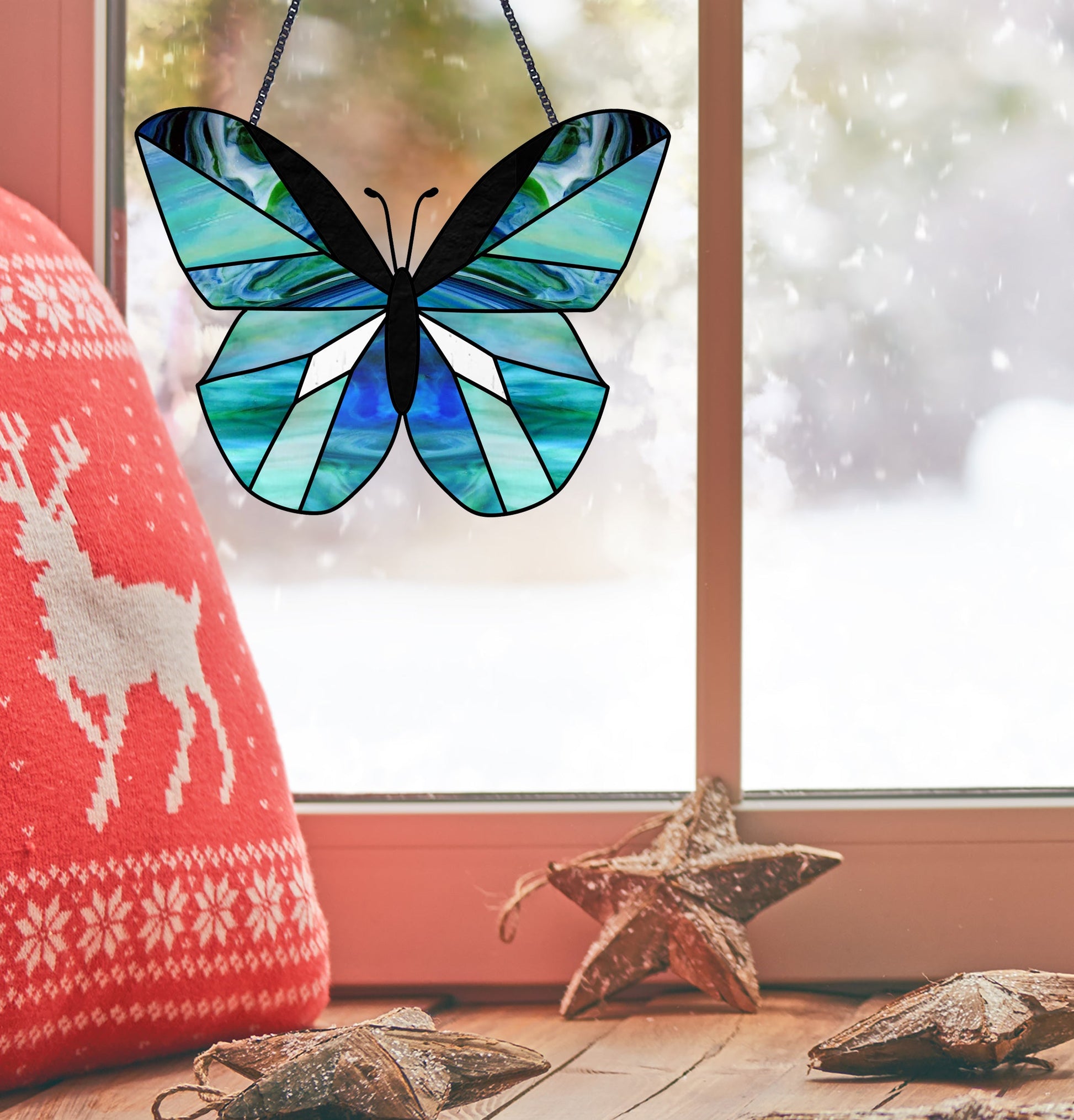 A stained glass butterfly created from the Beginner Butterfly Stained Glass Pattern in blue and teal hues hangs before a snowy window. A red cushion with a white reindeer sits on the left, while two wooden star ornaments adorn the windowsill as snow falls outside.