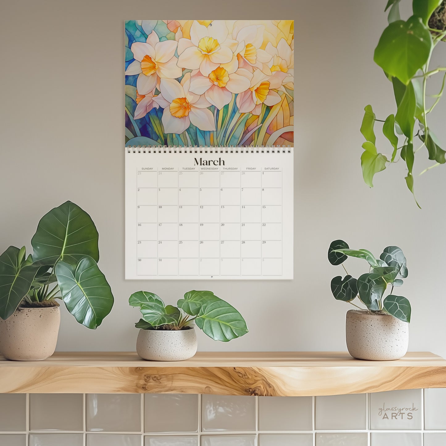 The March page of the 2025 Birth Flowers Wall Calendar displays vibrant daffodils above three potted plants on a wooden shelf, set against a light gray wall. A hint of stained glass floral illustrations can be seen in the hanging plant at the top right corner.