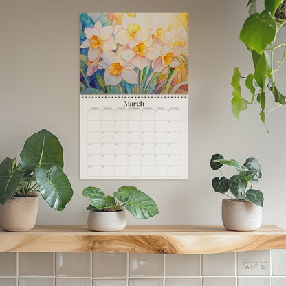 The birth flowers calendar, a 2025 monthly wall calendar with floral-themed original artwork, hanging on a wall surrounded by plants. The March page shows colorful daffodils.