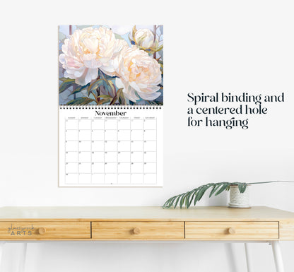 The 2025 Birth Flowers Wall Calendar presents Novembers floral artwork of white peonies in a stained glass style above the dates, complete with spiral binding and a hanging hole. It sits on a wooden desk next to a small plant against a plain white backdrop.