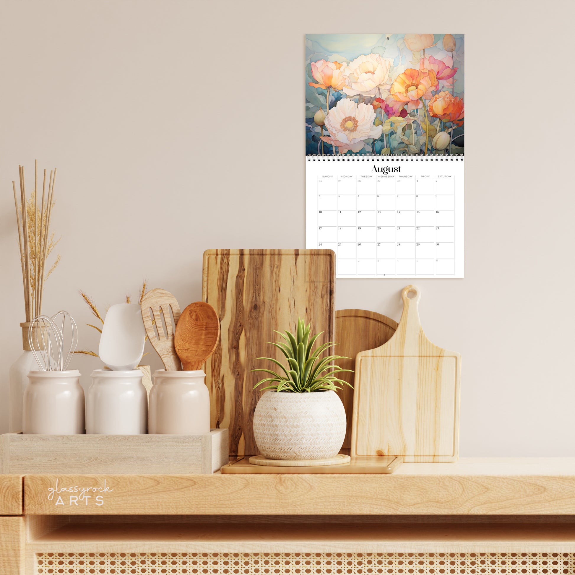 A kitchen counter with wooden utensils, a small potted plant, and cutting boards is set against a neutral-toned wall, complemented by a vibrant 2025 Birth Flowers Wall Calendar featuring stunning stained glass floral artwork for August.