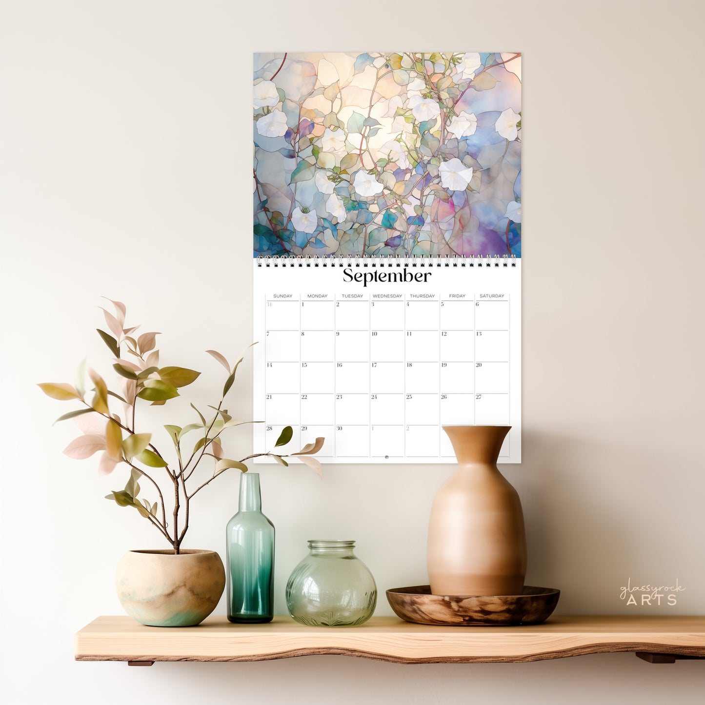 A 2025 Birth Flowers Wall Calendar with stained glass floral illustrations for September adorns the wall above a wooden shelf. Ceramic vases, glass bottles, and a small plant grace the shelf, their colors harmonizing with the light background to enhance the artistic display.