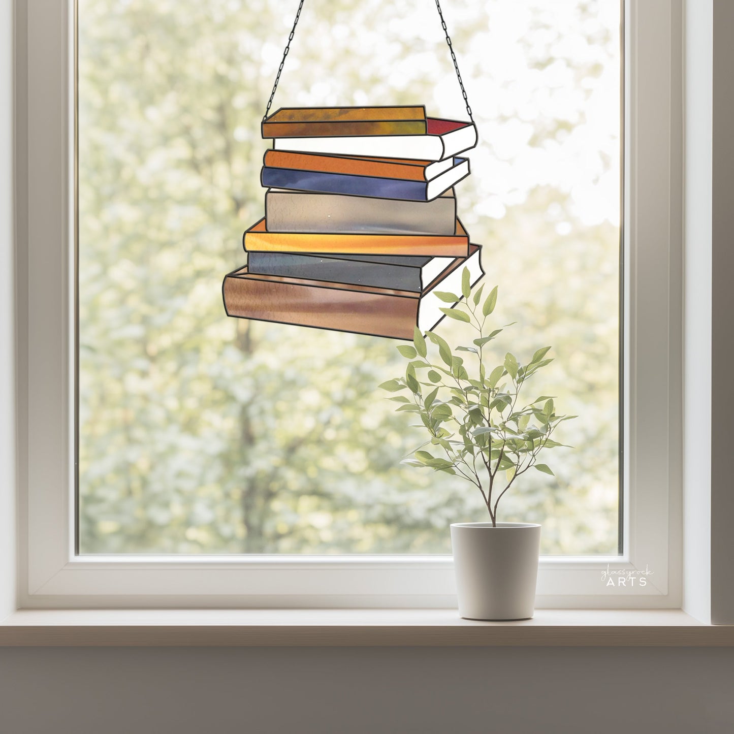 The Books Stained Glass Pattern, ideal as a gift for book lovers, hangs near a window resembling stacked books. Outside, blurred green foliage sets the backdrop while a small potted plant on the windowsill adds a natural touch to the scene.