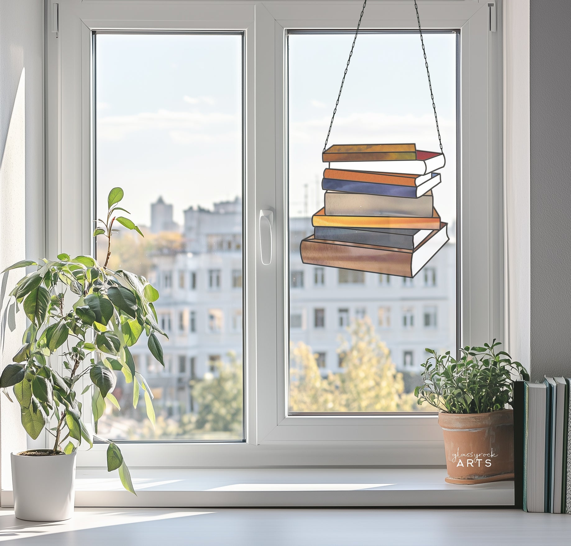 A window reveals city buildings and trees, adorned with the hanging Books Stained Glass Pattern, perfect as a book lovers gift. A houseplant sits gracefully on the windowsill beside herbs and some cherished reads.