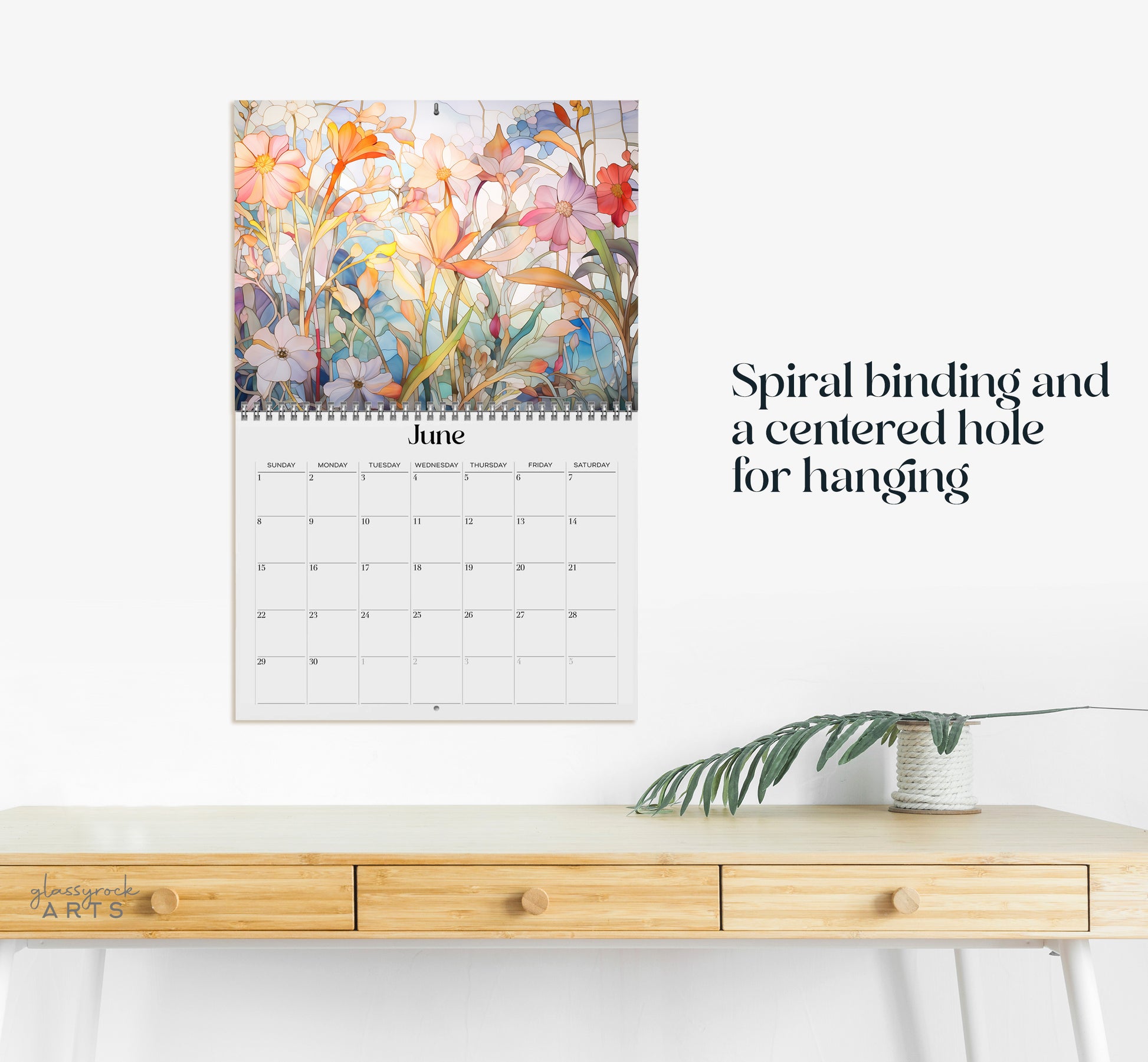 The 2025 Wall Calendar - Stained Glass Tropical Blooms, showcasing the month of June with spiral binding and a centered hole for hanging, features vivid tropical flower illustrations. It is positioned above a wooden desk adorned with a small potted plant.