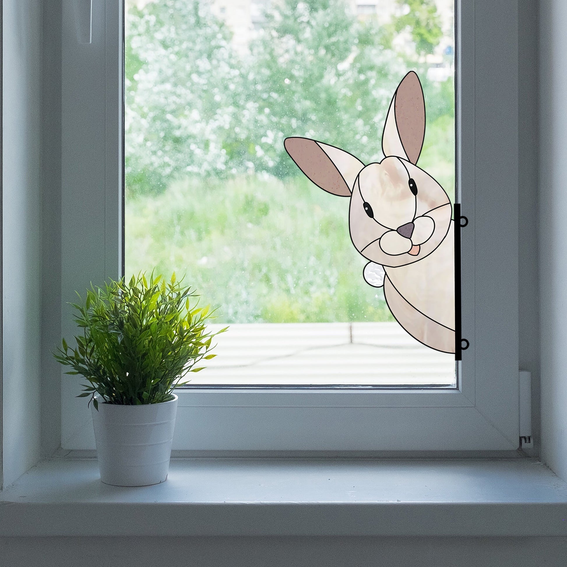 A cartoon bunny peeks from the right side of a white-framed window next to a potted plant, mirroring the Rabbit Buddy Bunny Stained Glass Rabbit Pattern. The view outside blends into a blurred green landscape.