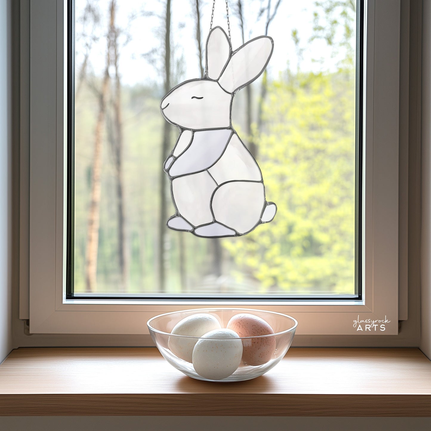 Hanging on the window with a view of trees, the Bunny Stained Glass Pattern decoration catches sunlight. Below, a clear bowl on the sill holds three eggs in different hues.