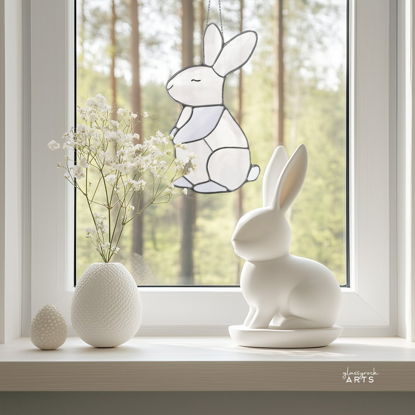 A white ceramic rabbit and an egg-shaped ornament rest on a window sill with a textured vase as the Bunny Stained Glass Pattern sun catcher hangs in the window, casting colorful hues across the room against the forest view.