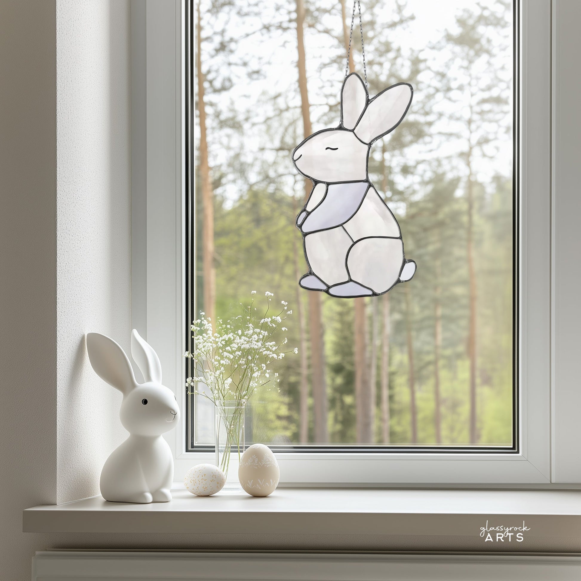 A minimalist interior features a windowsill with a white rabbit figurine, small white floral vase, and two decorative eggs. The Bunny Stained Glass Pattern hangs on the window with blurry green trees visible outside, ideal for digital download enthusiasts.