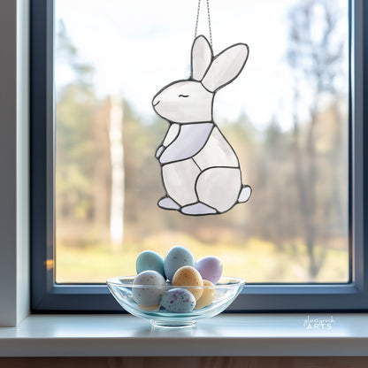 A Bunny Stained Glass Pattern catches the light above a windowsill, where a glass bowl filled with pastel eggs rests. Beyond the window, softly blurred trees create a captivating backdrop for this charming sun catcher.