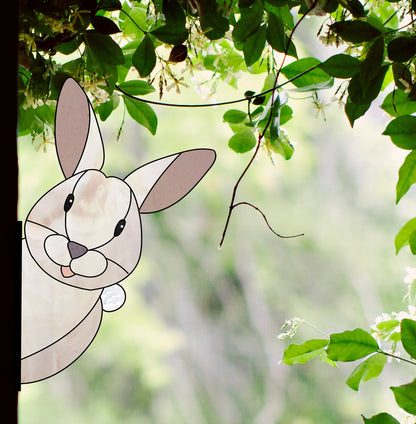 Illustration of a cartoon bunny peeking around a window frame amid lush greenery and blurred foliage. This Rabbit Buddy Bunny Stained Glass Rabbit Pattern adds whimsical charm to any space.