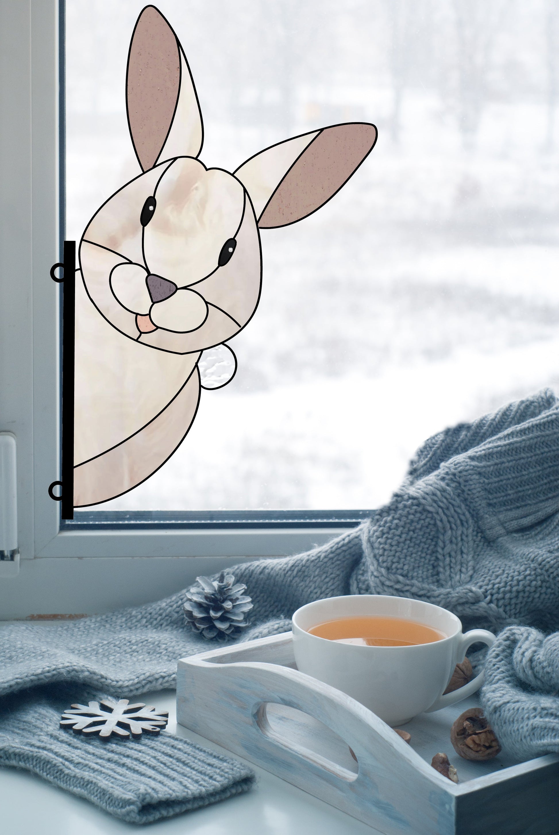 A window showcases the Rabbit Buddy Bunny Stained Glass Rabbit Pattern, its smile glowing. Beyond lies a snowy landscape. Inside, a cozy scene with a knit blanket, wooden tray with tea, pinecones, and snowflake decor offers ideal inspiration for digital pattern downloads.