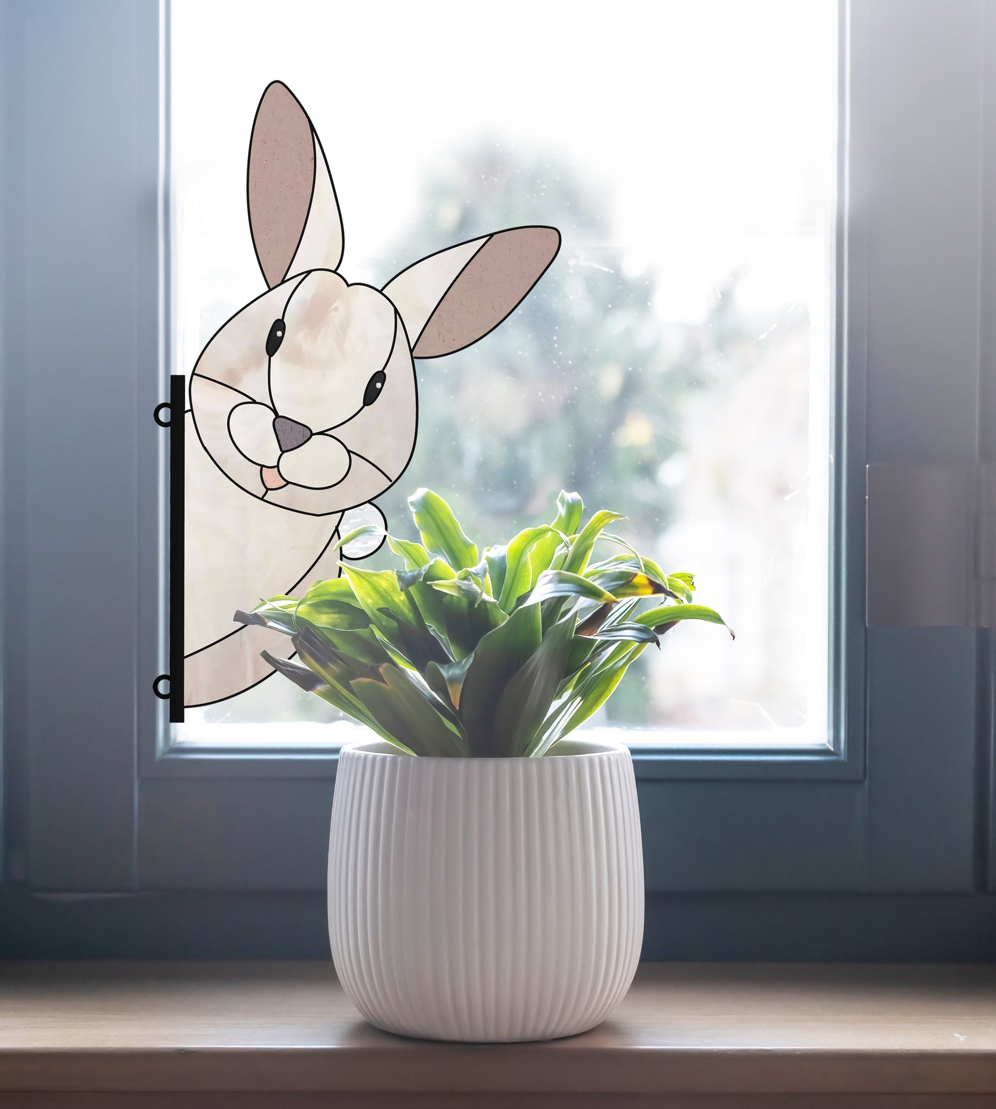 A potted plant with green leaves sits on a windowsill beside the Rabbit Buddy Bunny Stained Glass Rabbit Pattern, showing a curious rabbit peeking inside against a blurred outdoor view.