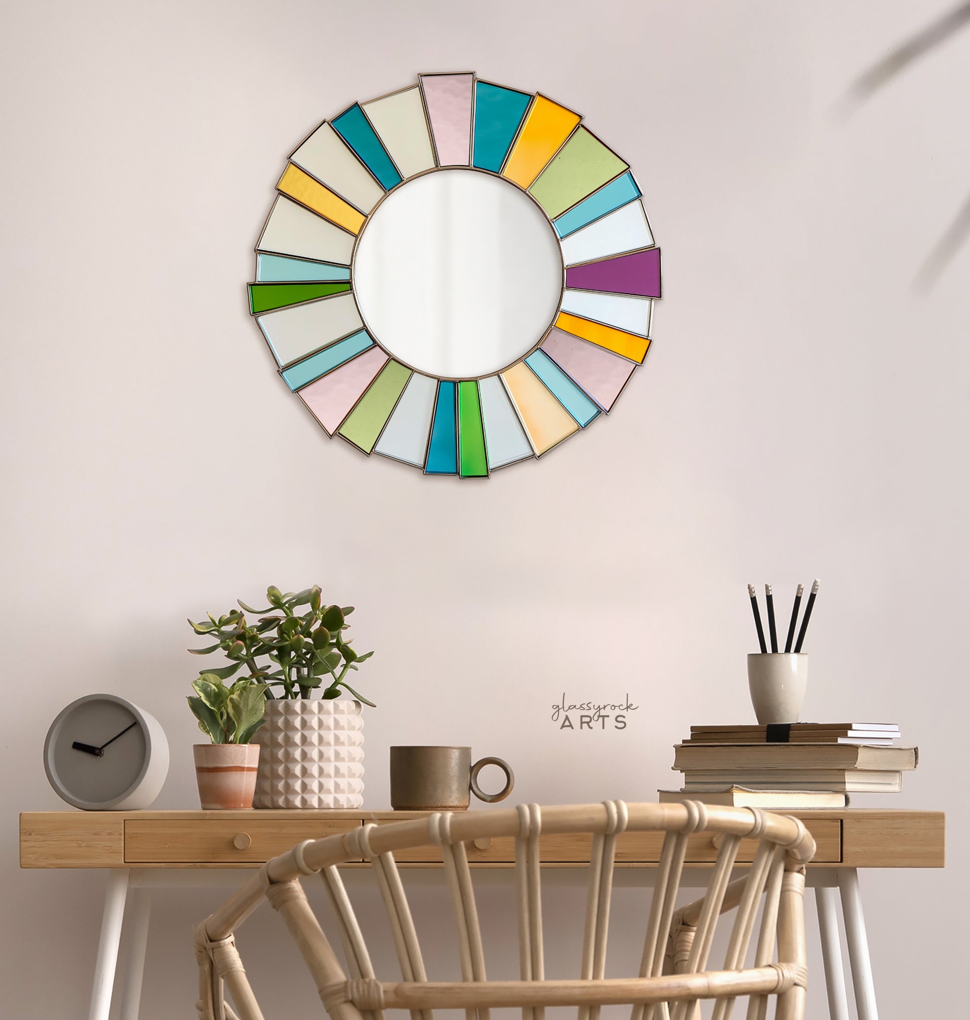 A round mirror surrounded by bright multicolored pieces of stained glass radiating from the edges of the mirror. It hangs on a wall above a desk with boho items.