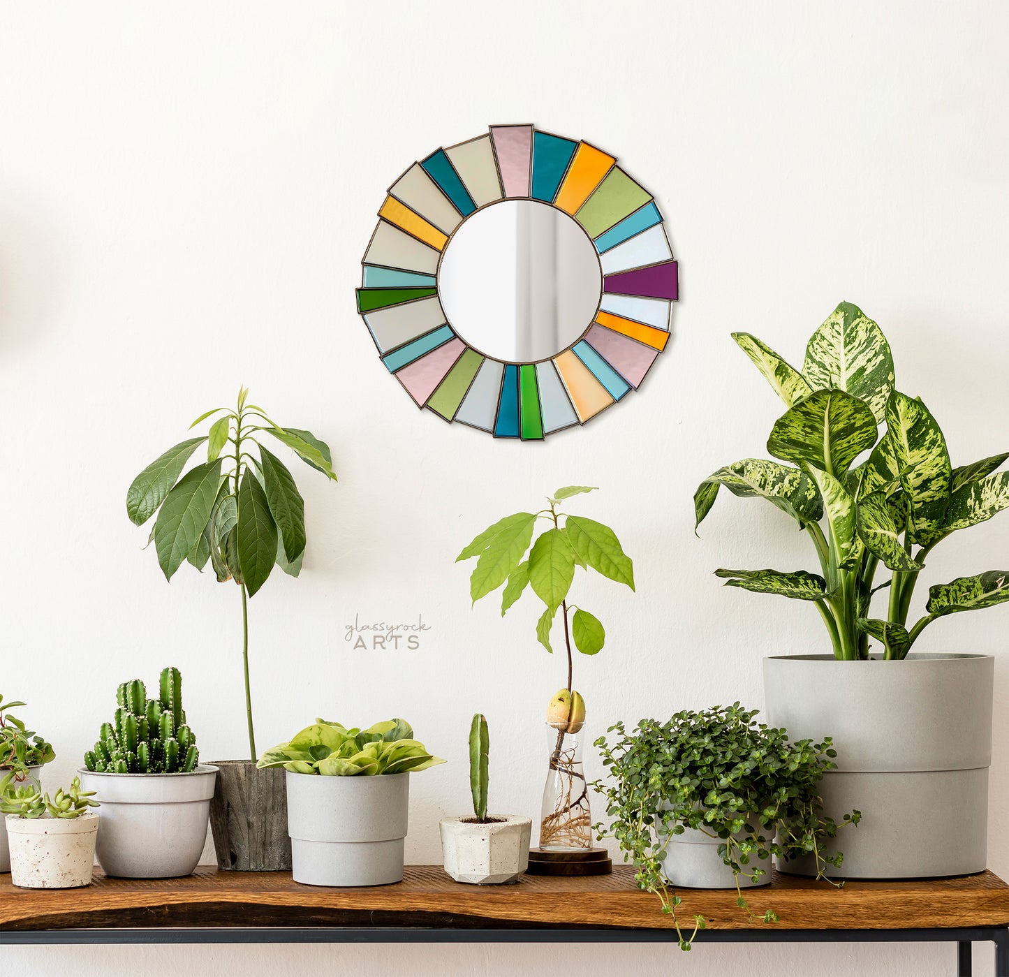 A round mirror surrounded by bright multicolored pieces of stained glass radiating from the edges of the mirror. It hangs on a wall with a table full of plants below.