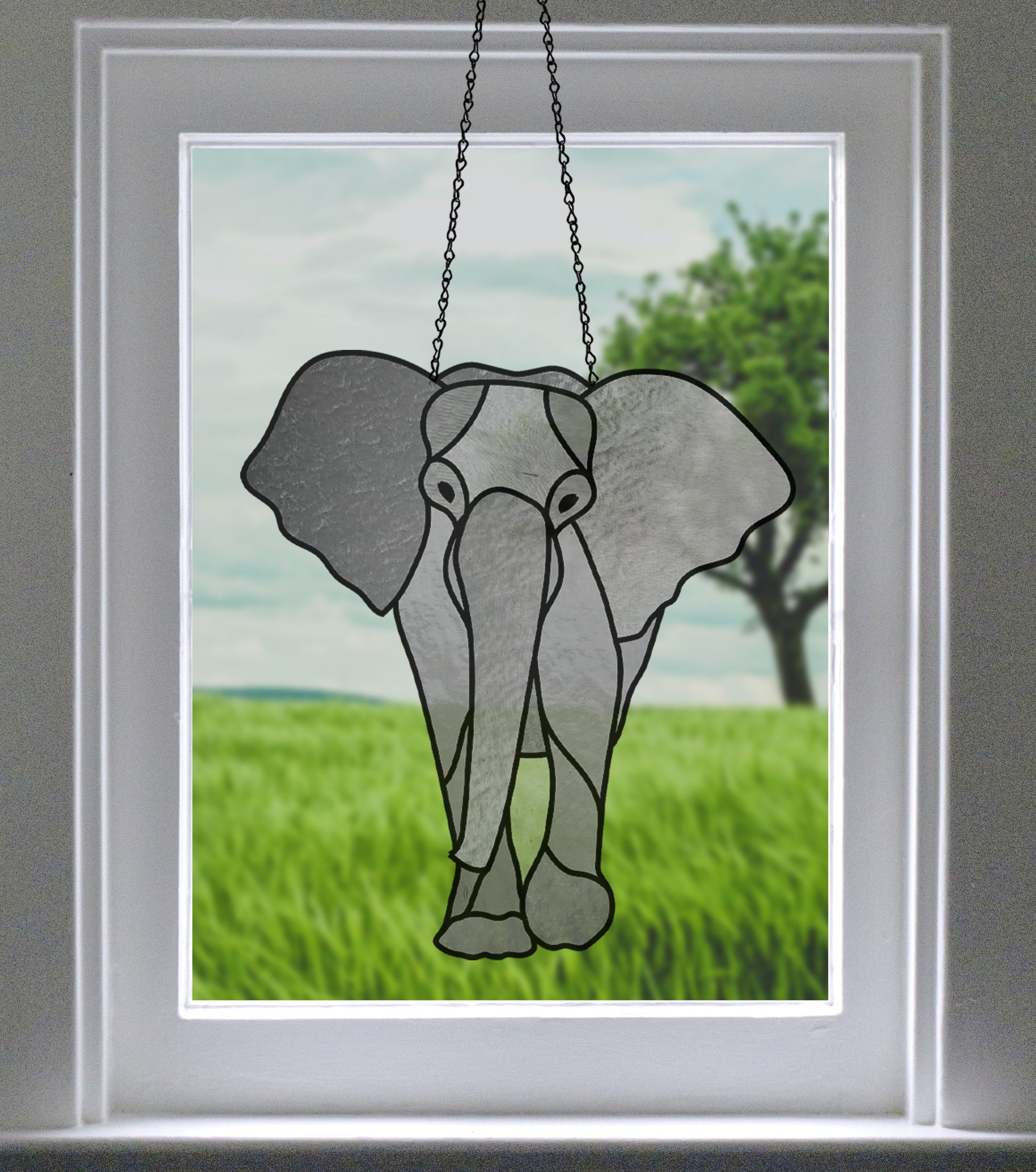 A stained glass elephant head decoration hangs in front of a window with a view of a green field and a tree in the background.