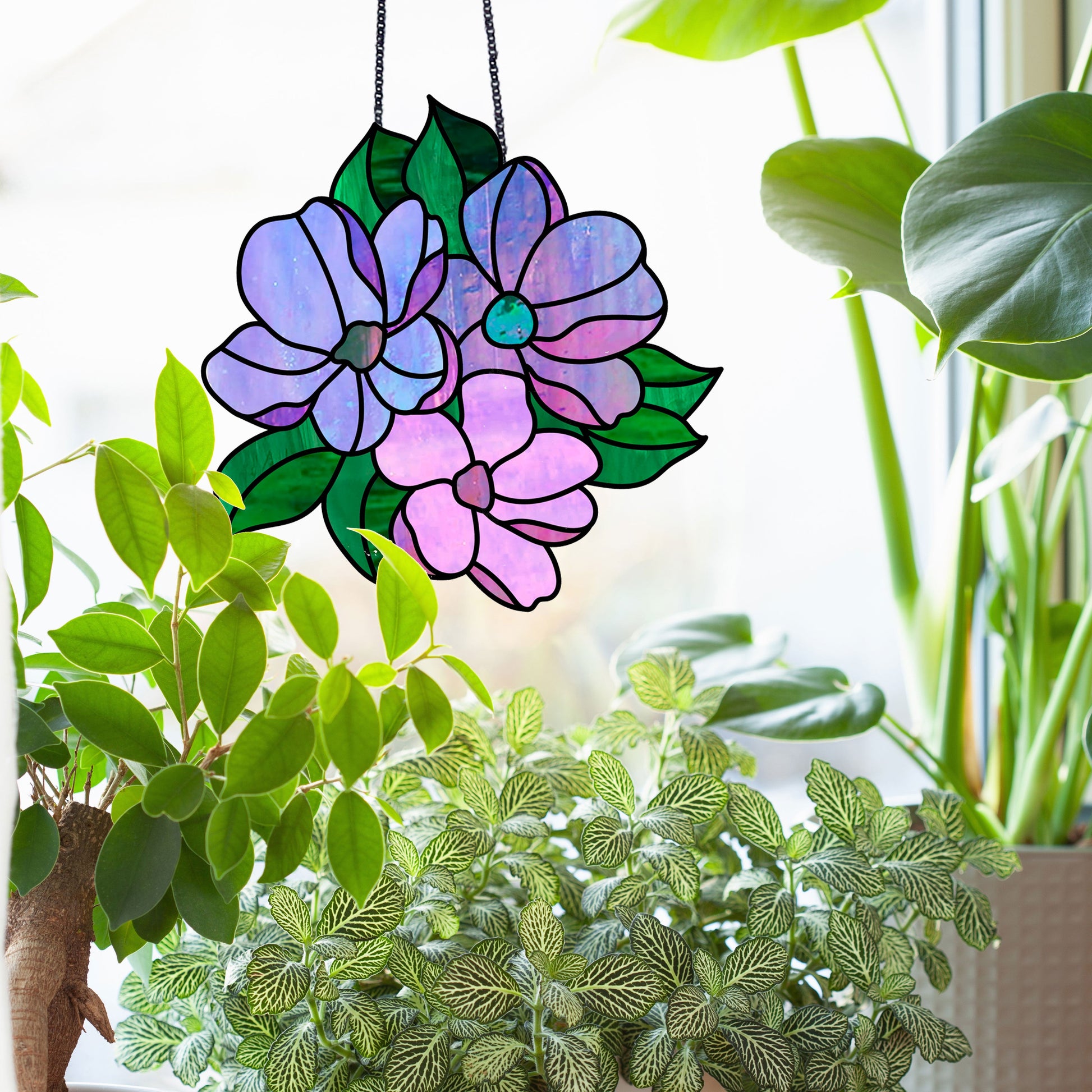 The Bundle of Flowers Stained Glass Pattern suncatcher, featuring purple blooms and green leaves, gracefully hangs in front of a window. Surrounded by lush indoor plants, it transforms natural light into a vibrant display.